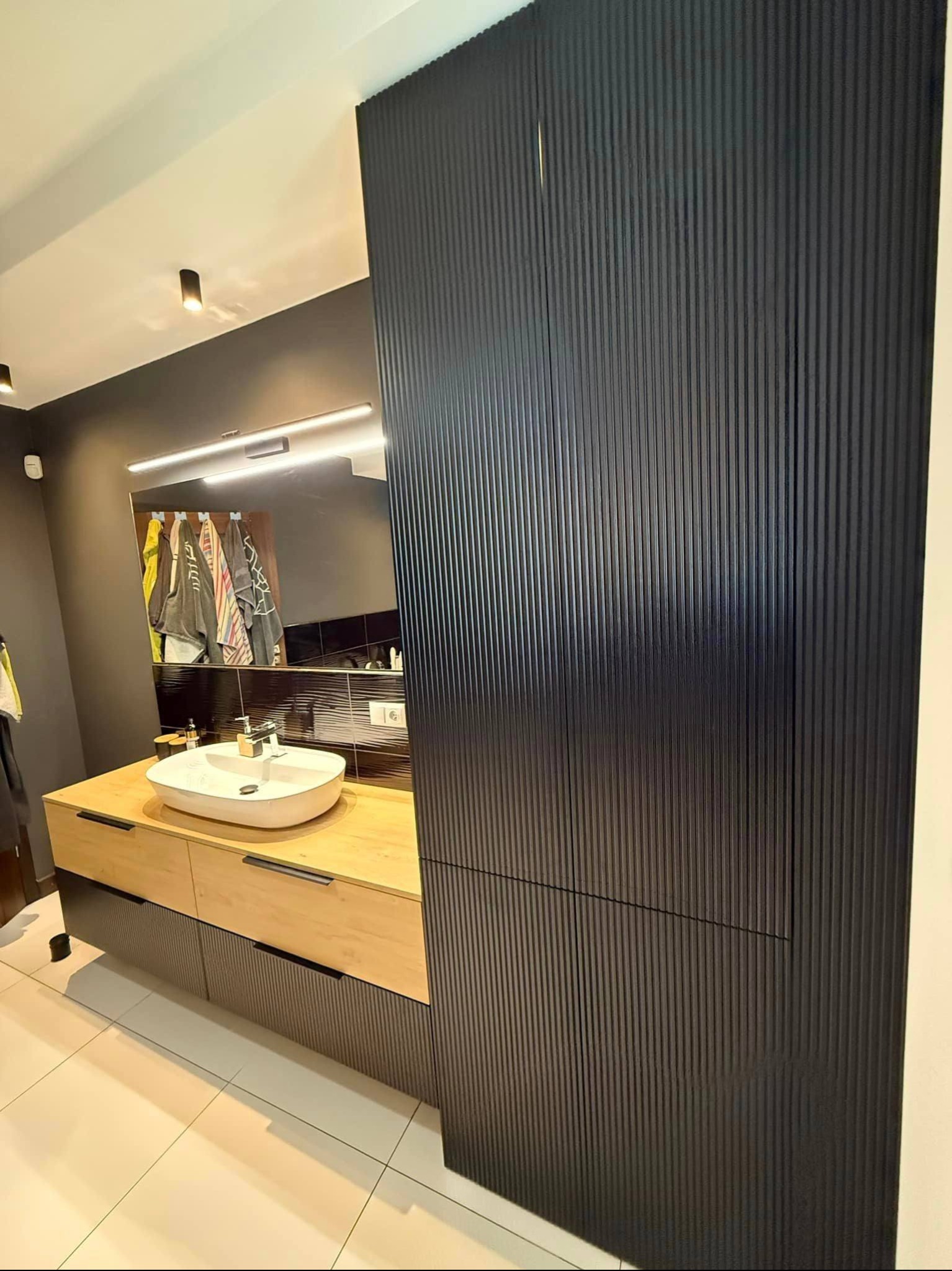 Nowoczesna łazienka z umywalką nablatową, drewnianą szafką i wysoką, czarną szafą z pionowymi żłobieniami. Lustro z oświetleniem LED.