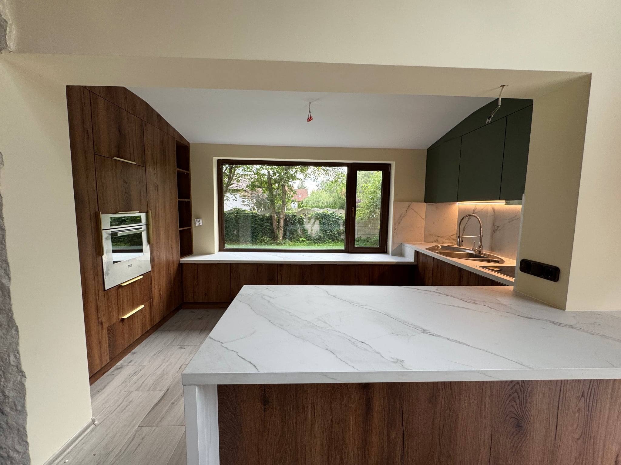 Nowoczesna kuchnia z wyspą i drewnianymi frontami, marmurowym blatem, oświetleniem LED i widokiem na ogród przez okno.