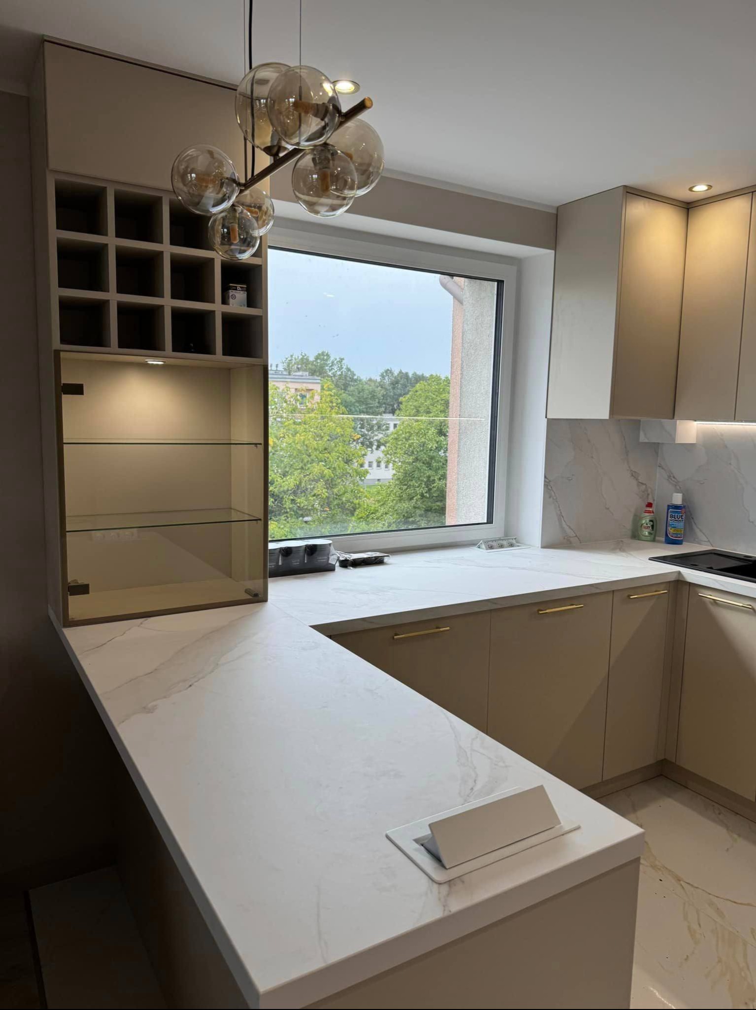 Beżowa kuchnia z blatem imitującym marmur, przeszklona witryna z półkami i moduł na wino. Nad blatem okno z widokiem na zieleń. Nowoczesne oświetlenie.