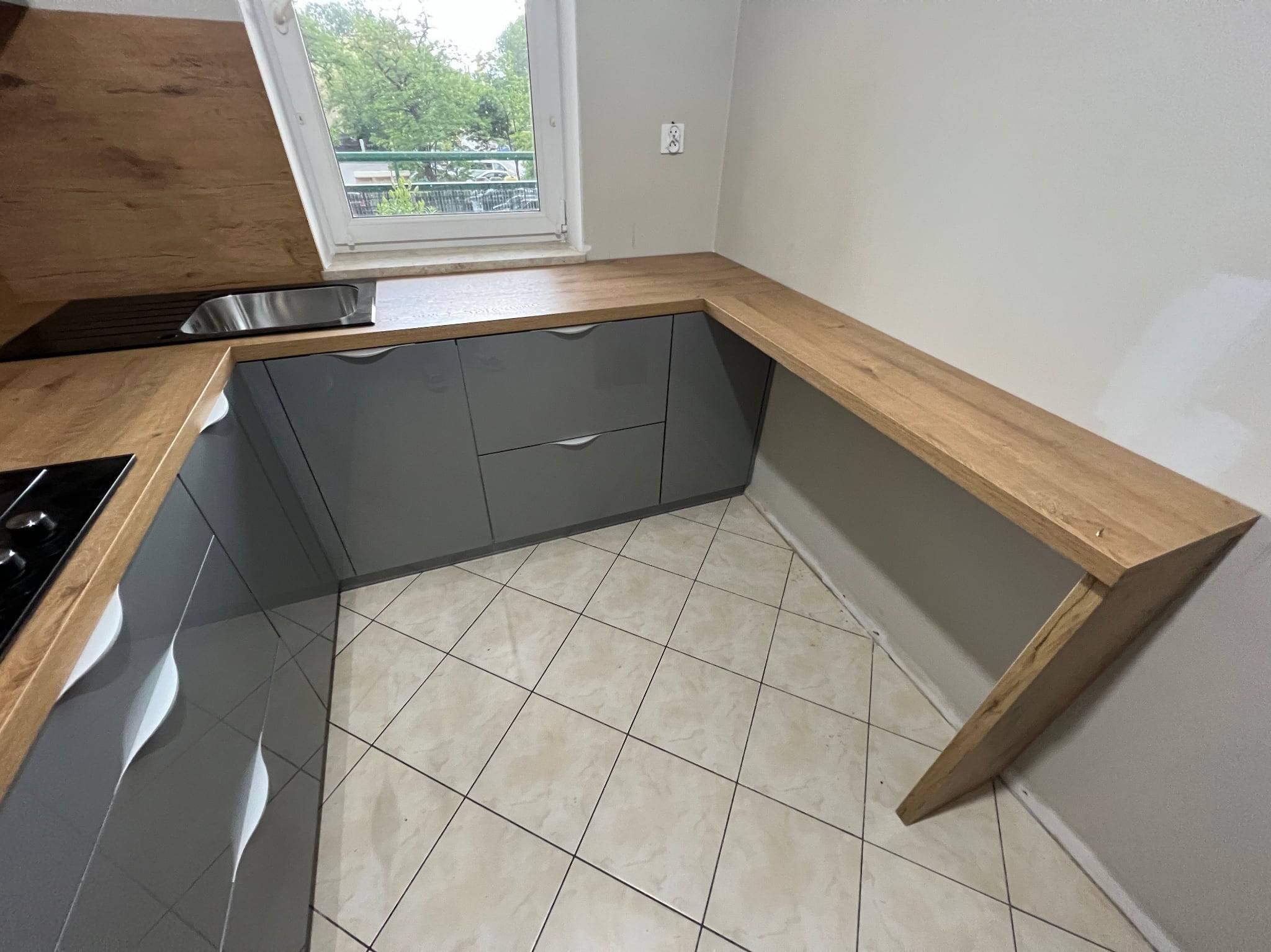 Szare szafki kuchenne z drewnianym blatem przy oknie. Widoczny zlew i płyta grzewcza. Jasna podłoga z płytek. Oświetlenie naturalne z okna.