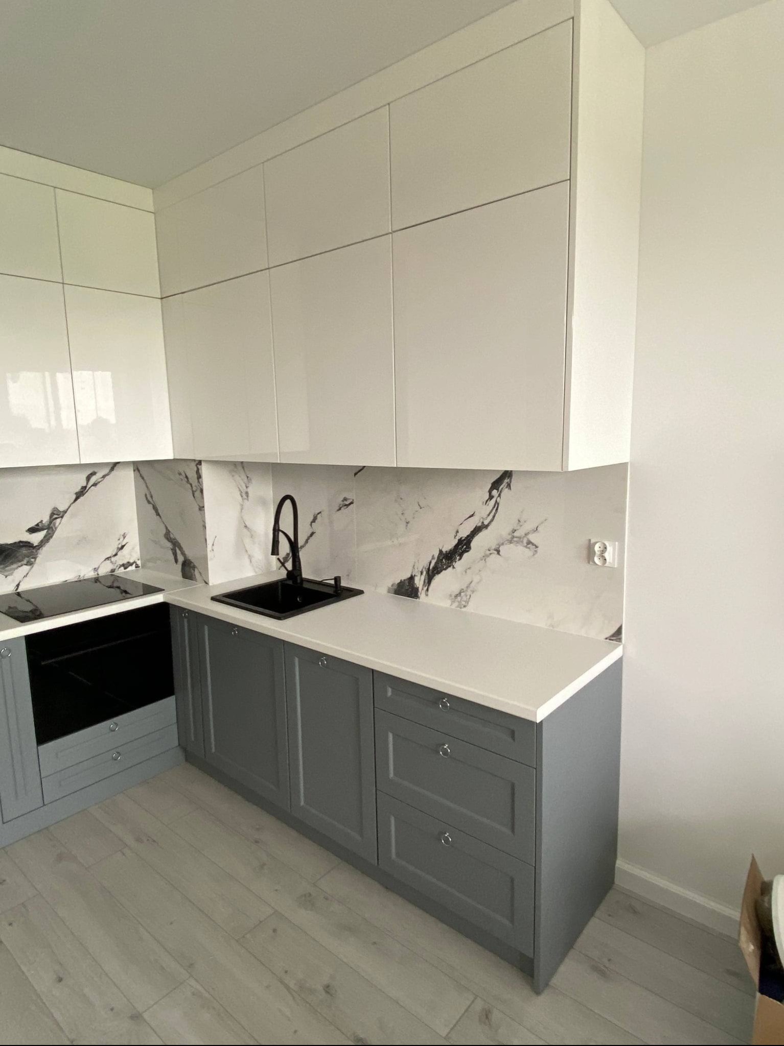 Szara zabudowa kuchenna z białymi szafkami górnymi i marmurowym backsplashiem. Czarna armatura i płyta indukcyjna. Jasna podłoga. Minimalistyczny styl.