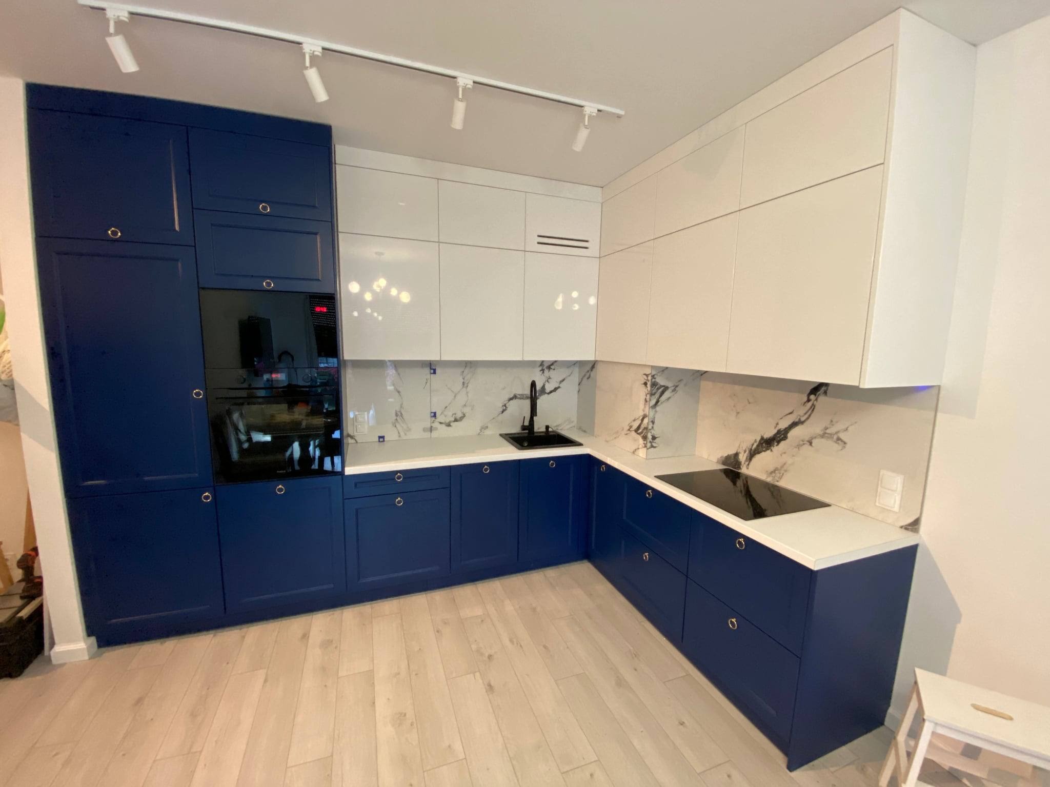 Nowoczesna kuchnia: granatowe dolne szafki z okrągłymi uchwytami, białe górne szafki, marmurowe płytki na ścianie. Oświetlenie punktowe na suficie.