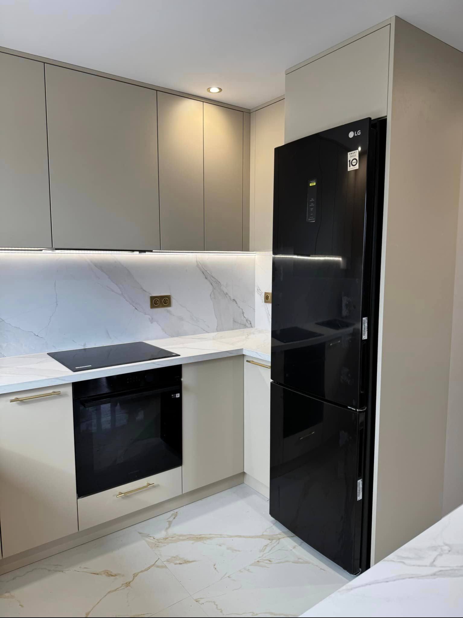 Beżowa kuchnia z czarną lodówką LG, blatem imitującym marmur i złotymi uchwytami. Oświetlenie LED pod szafkami. Nowoczesny design.