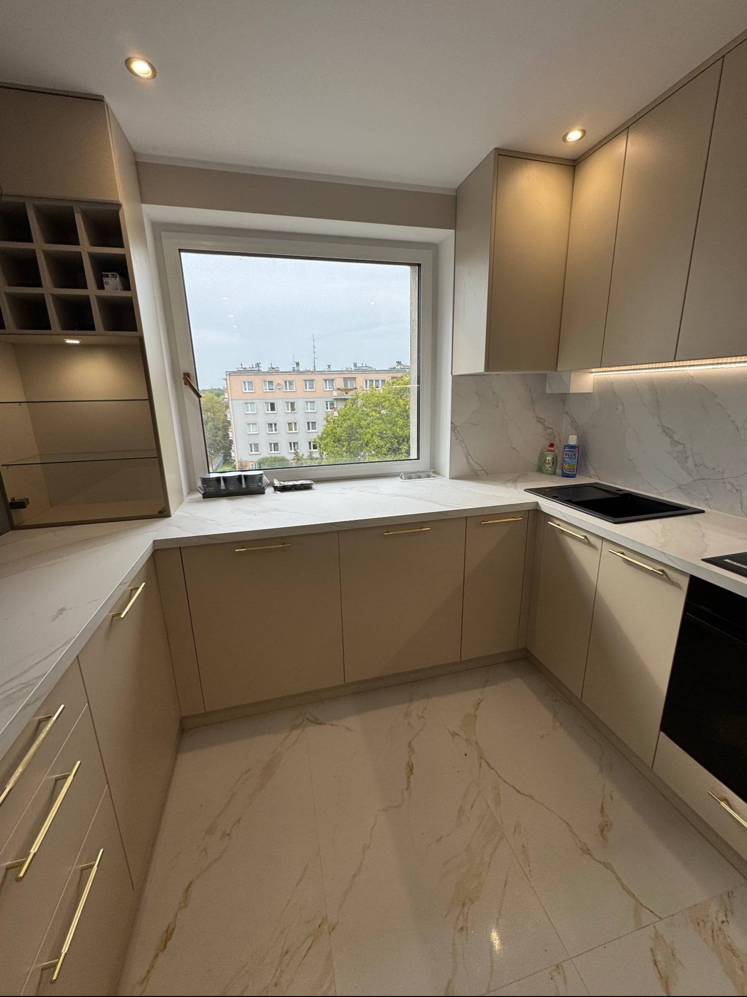 Beżowa kuchnia z marmurowym blatem i złotymi uchwytami, widok na miasto przez okno. Nowoczesne oświetlenie podszafkowe, minimalistyczny design.