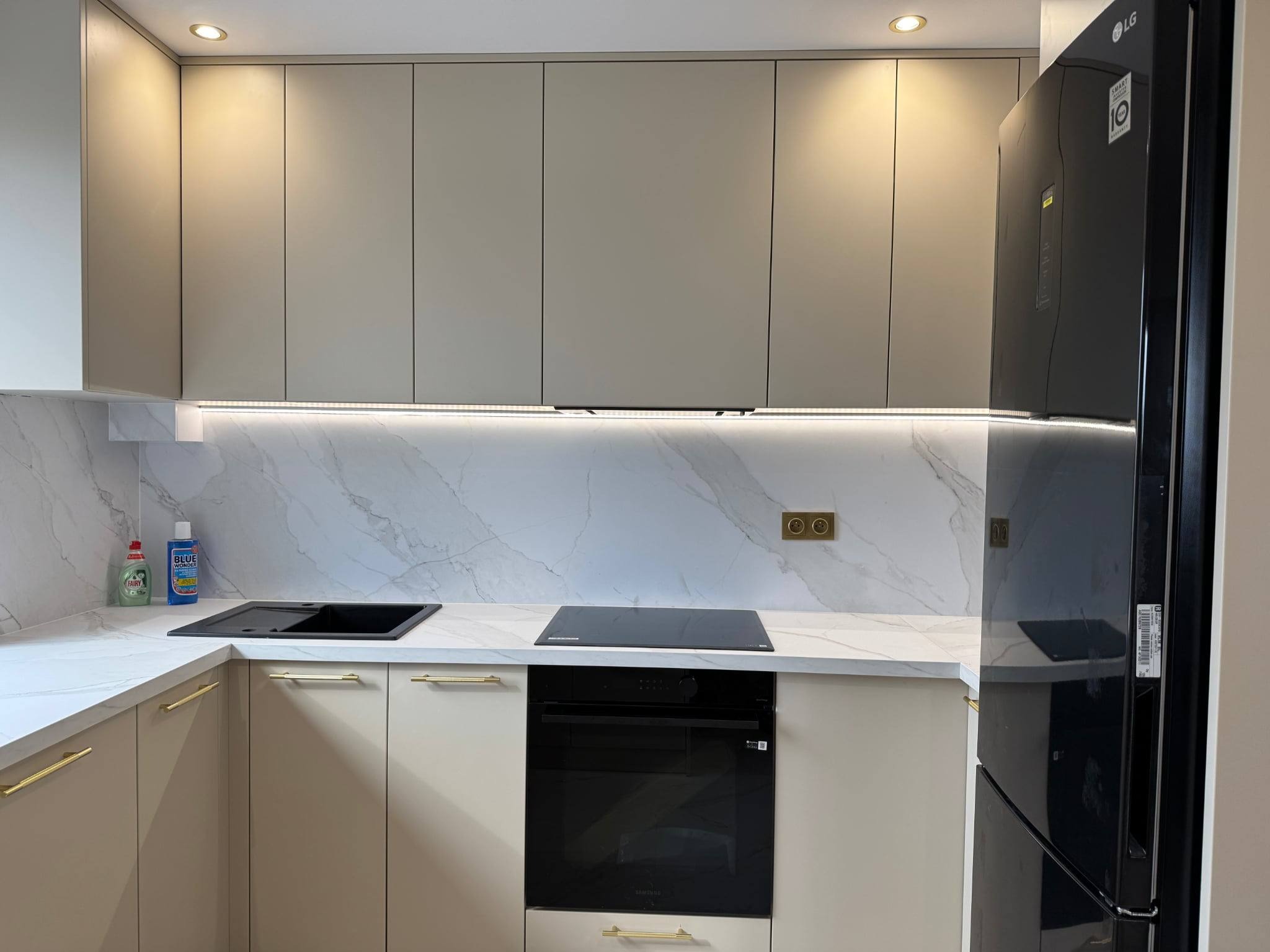 Beżowa kuchnia na wymiar z marmurowym blatem i złotymi uchwytami. Widoczna czarna lodówka LG, płyta indukcyjna i piekarnik. Podświetlenie LED pod szafkami.