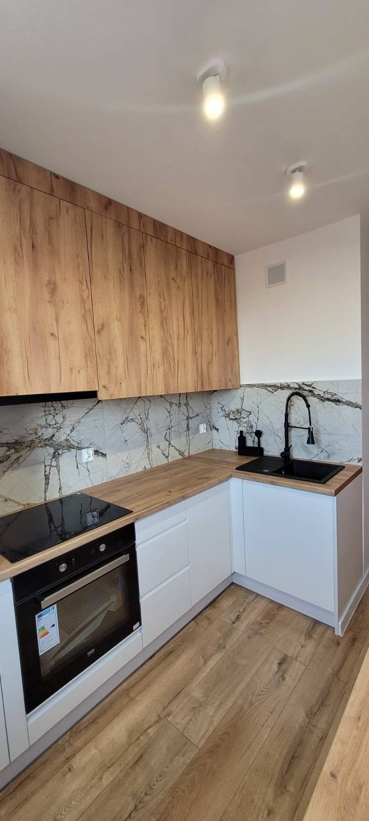 Nowoczesna kuchnia w Radomsku: białe szafki dolne, drewnopodobne górne, blat i podłoga. Czarny zlew i bateria, marmurowy backsplash, oświetlenie punktowe.