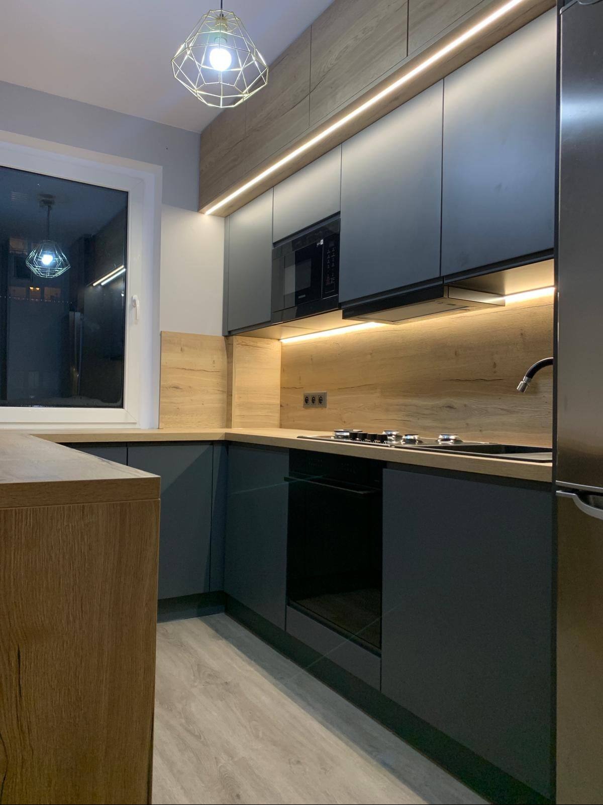 Nowoczesna kuchnia z szarymi frontami i drewnianym blatem, oświetlona ledami. Widoczny piekarnik, płyta grzewcza i okno z odbiciem lampy. Minimalistyczny design.