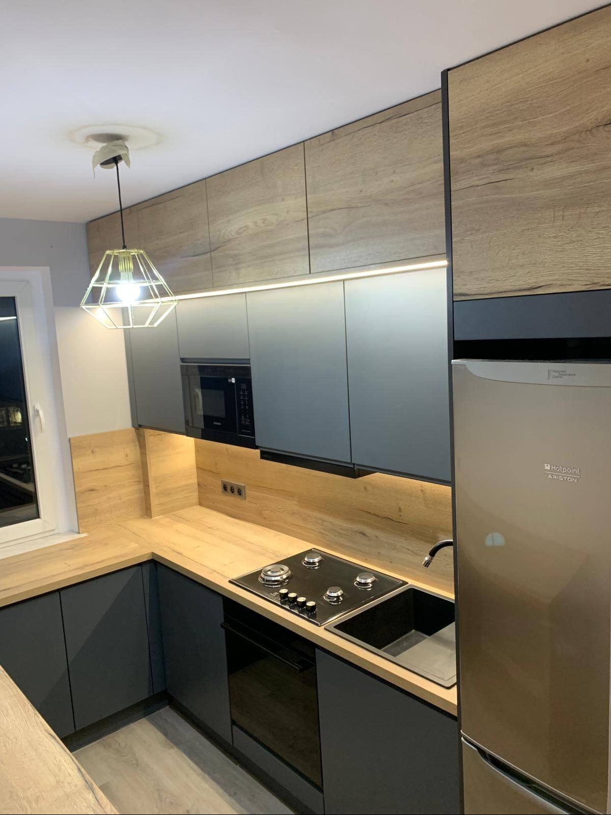 Nowoczesna kuchnia z szarymi frontami i drewnianymi akcentami, oświetlona listwą LED pod szafkami. Widoczny fragment lodówki i płyty grzewczej.