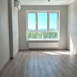Andrii Derevianko - Jasne, puste pomieszczenie z panelami podłogowymi, grzejnikiem pod oknem i klimatyzatorem na ścianie. Widok na zieleń za oknem. Żarówka zwisa z sufitu.