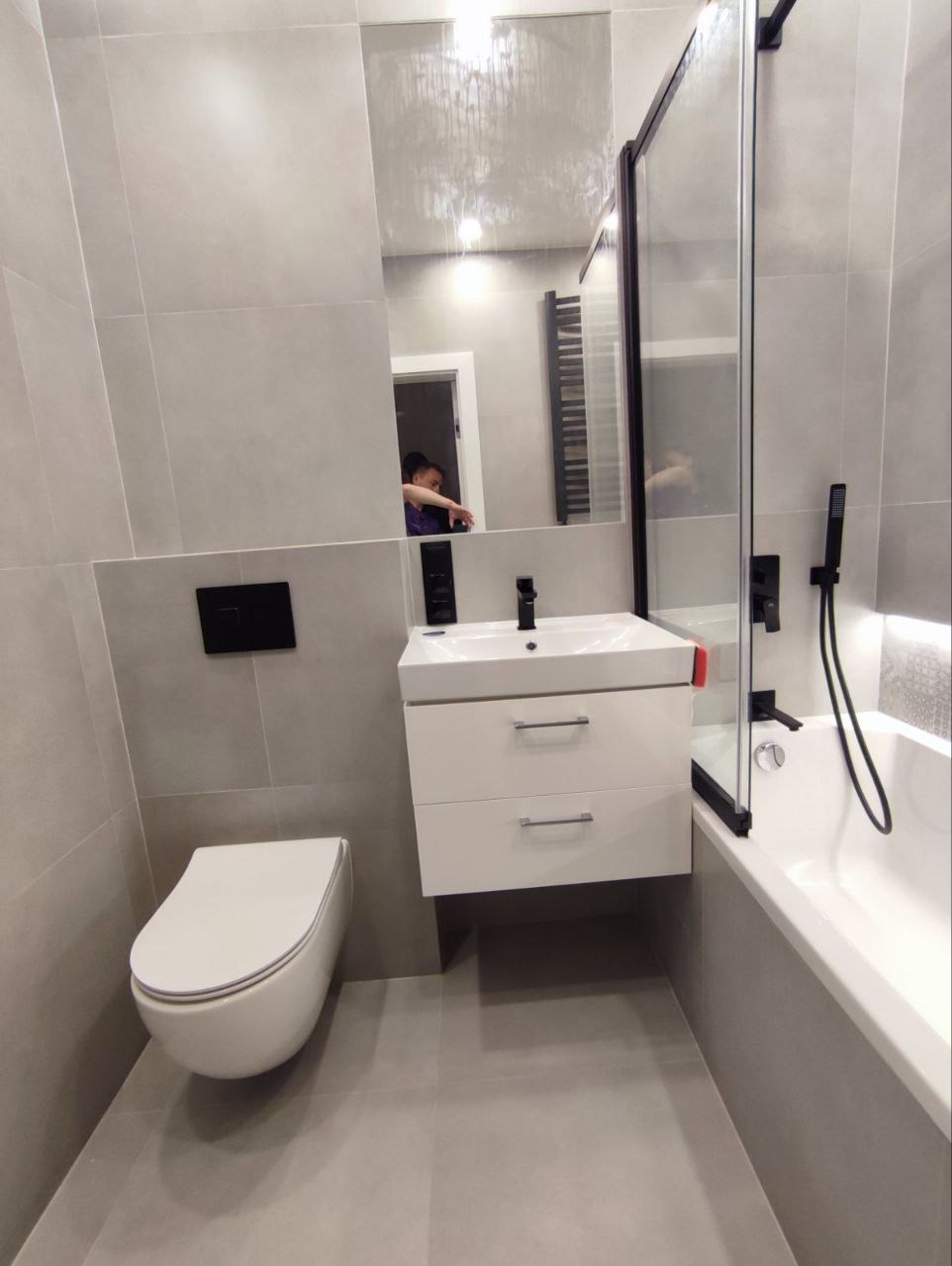 Nowoczesna łazienka w szarościach: podwieszana toaleta, biała szafka z umywalką, czarna armatura i kabina prysznicowa z lustrem. Minimalistyczny design.