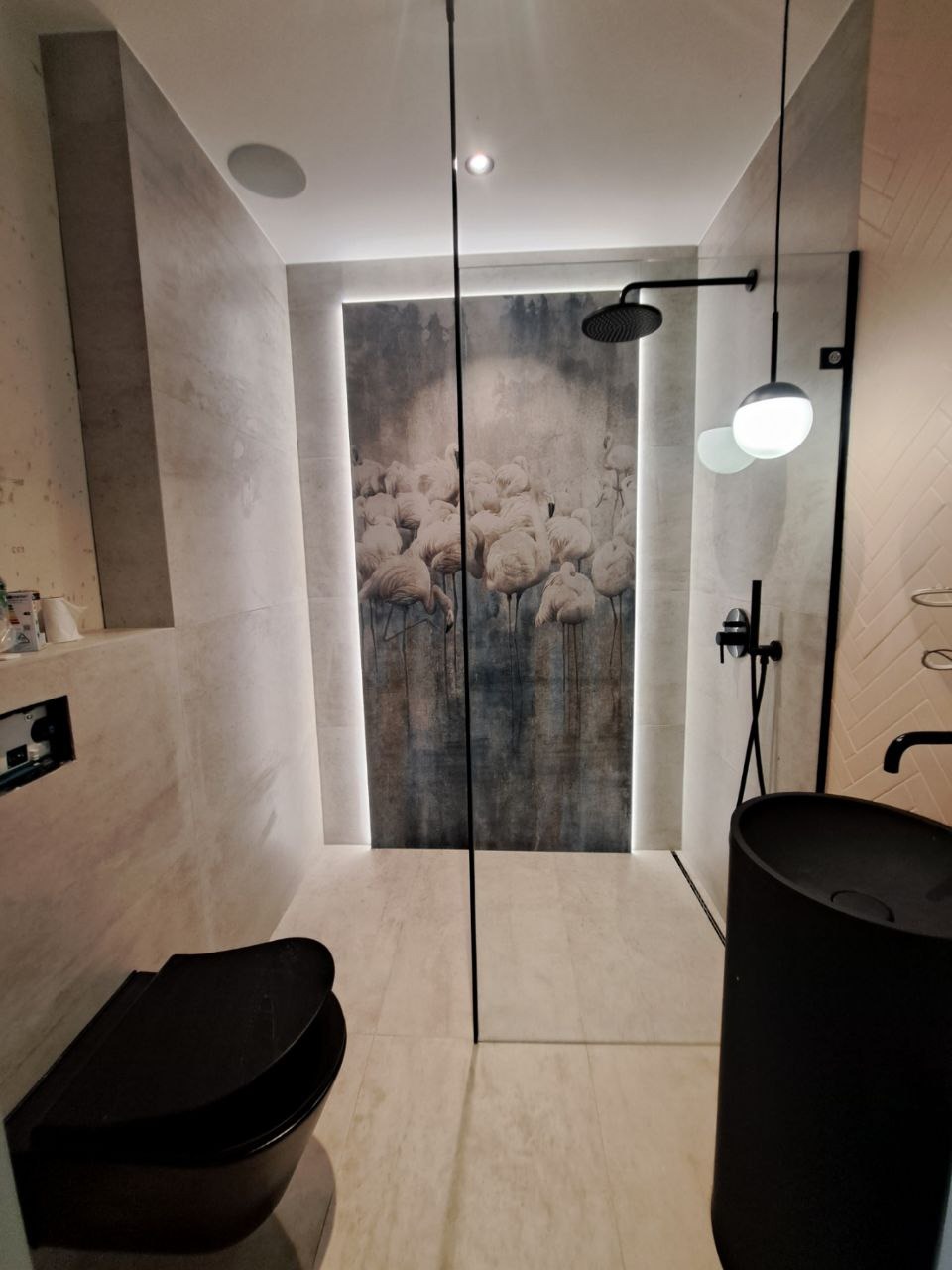 Nowoczesna łazienka z czarną armaturą, kabiną prysznicową typu walk-in i podświetlanym panelem dekoracyjnym z motywem flamingów. Minimalistyczny design, jasne płytki.