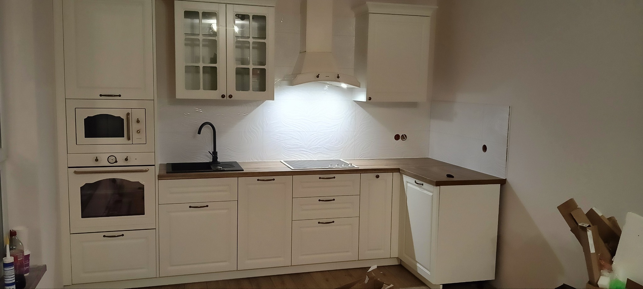 Jasna kuchnia w stylu klasycznym z drewnianym blatem, czarną armaturą i białymi płytkami z delikatnym wzorem. Widoczne zabudowane sprzęty AGD.