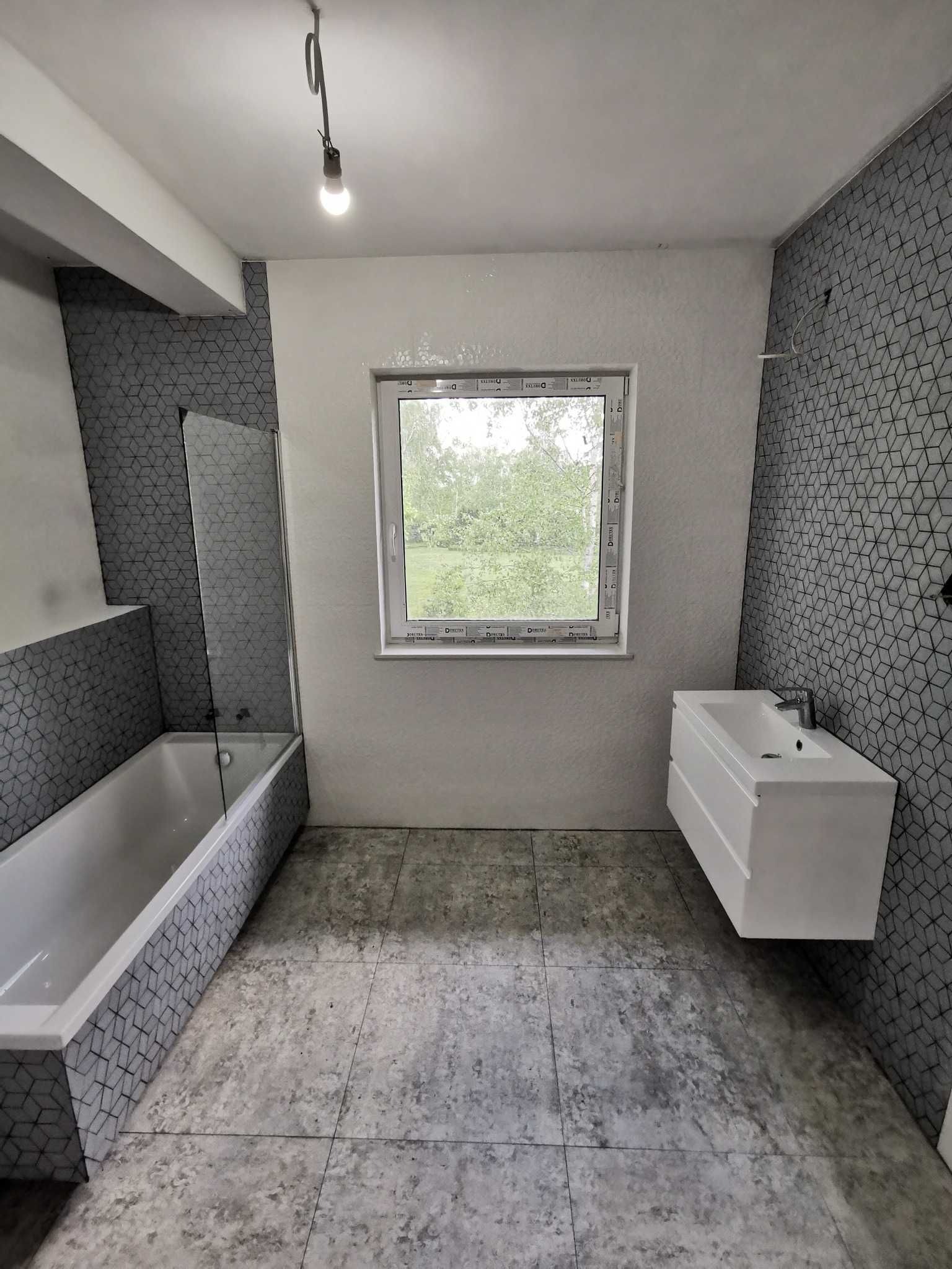 Wykończona łazienka z wanną, umywalką i oknem. Ściany i obudowa wanny wyłożone szarą mozaiką. Podłoga z szarych płytek imitujących beton. Minimalistyczny design.