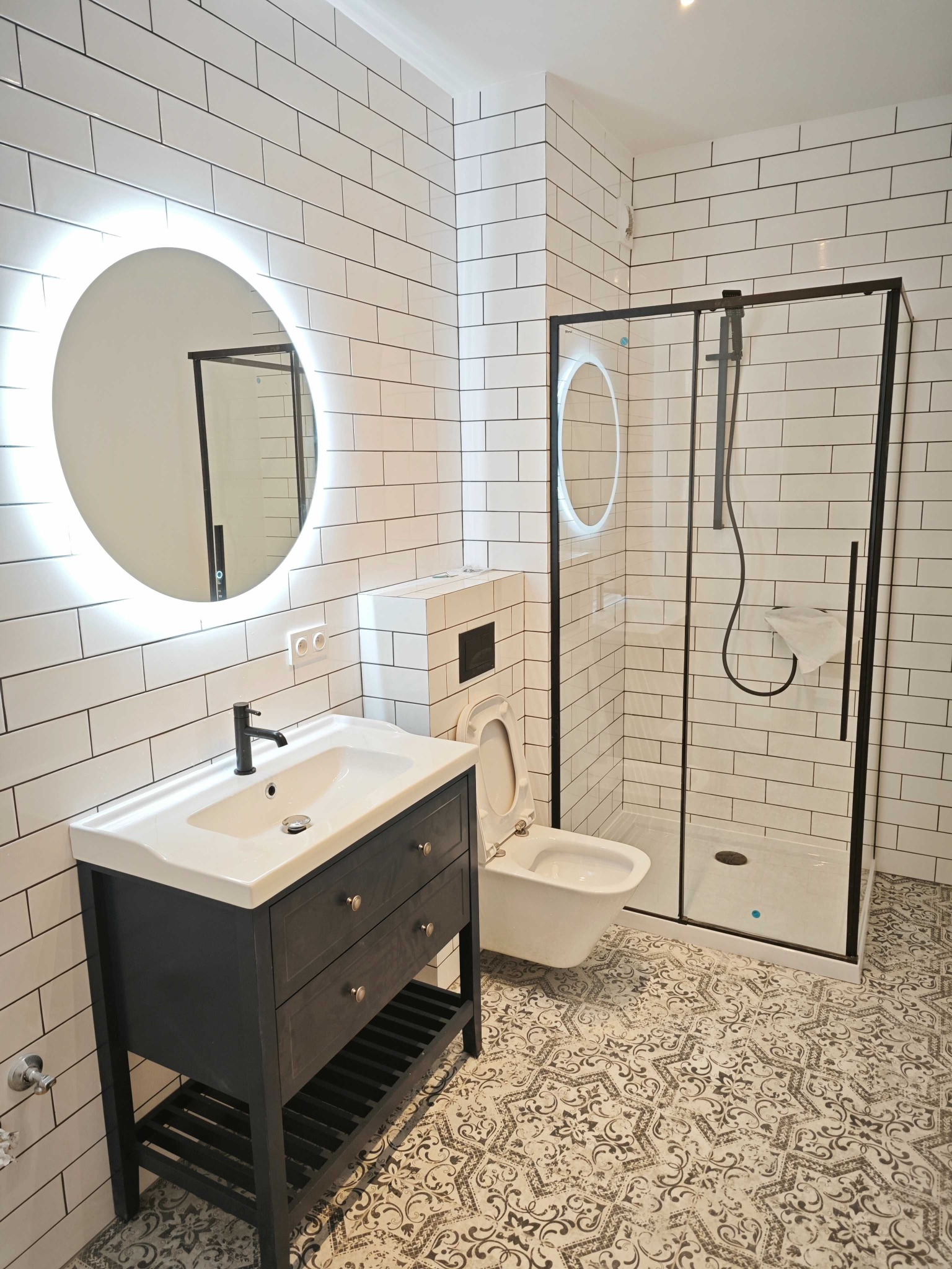 Nowoczesna łazienka z białymi płytkami, czarną armaturą i wzorzystą podłogą. Okrągłe lustro z podświetleniem LED nad umywalką. Kabina prysznicowa z czarną ramą.