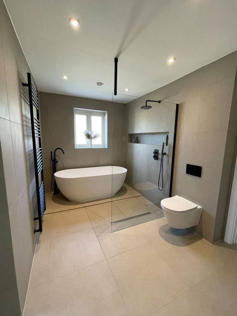 Nowoczesna łazienka z beżowymi płytkami, wolnostojącą wanną, kabiną prysznicową typu walk-in z czarnymi elementami i grzejnikiem drabinkowym.