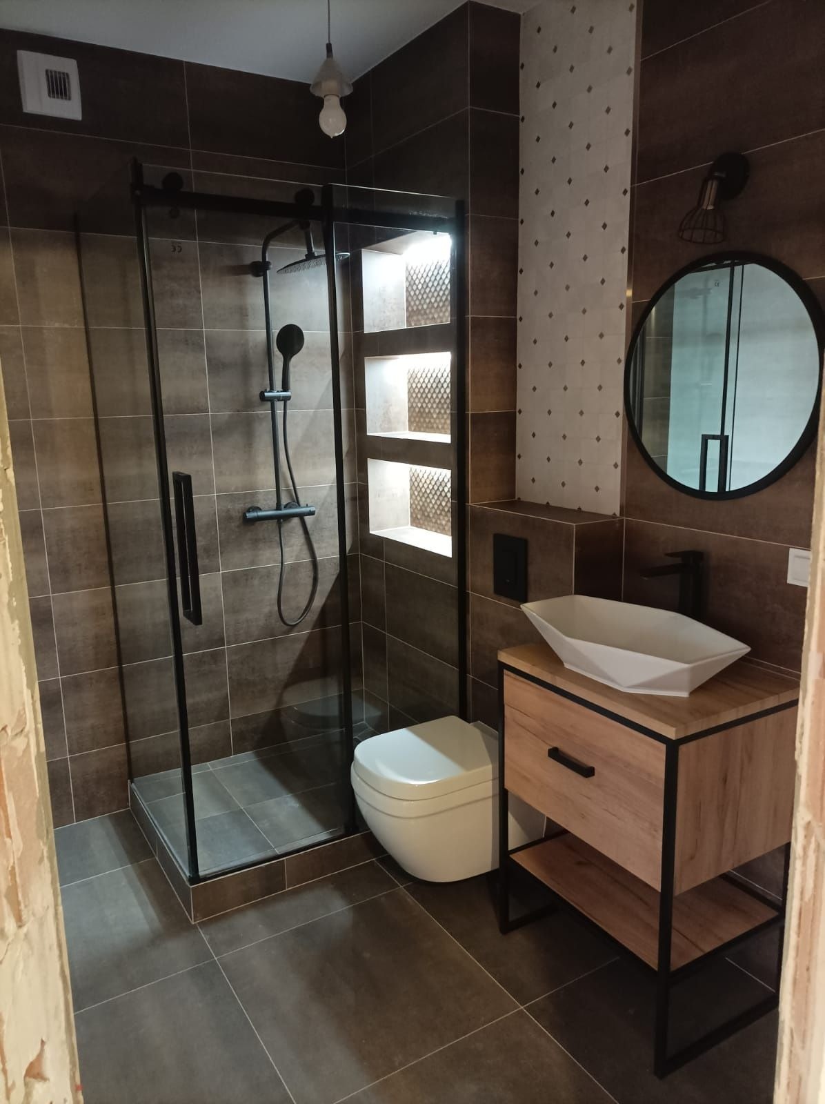 Nowoczesna łazienka z kabiną prysznicową typu walk-in, umywalką nablatową na szafce z drewnianym dekorem i okrągłym lustrem. Brązowe płytki i oświetlenie akcentujące.