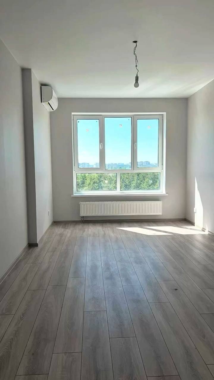 Jasne, puste pomieszczenie z panelami podłogowymi, grzejnikiem pod oknem i klimatyzatorem na ścianie. Widok na zieleń za oknem. Żarówka zwisa z sufitu.