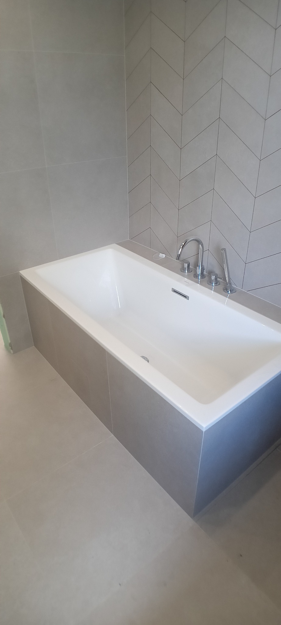 Nowoczesna, prostokątna wanna obudowana szarą płytką, z baterią i słuchawką. Na ścianie płytki ułożone w jodełkę. Minimalistyczna łazienka w szarościach.