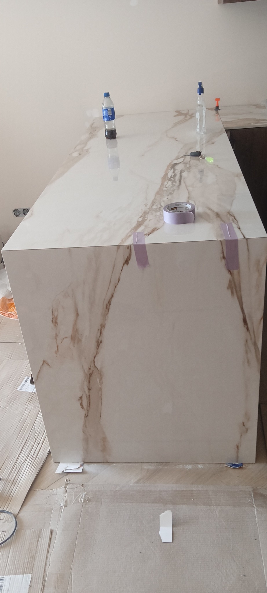 Wykończona wyspa kuchenna z płytek imitujących marmur, zabezpieczona taśmą malarską. Na blacie butelki i narzędzia glazurnicze. Widoczne ślady prac wykończeniowych.