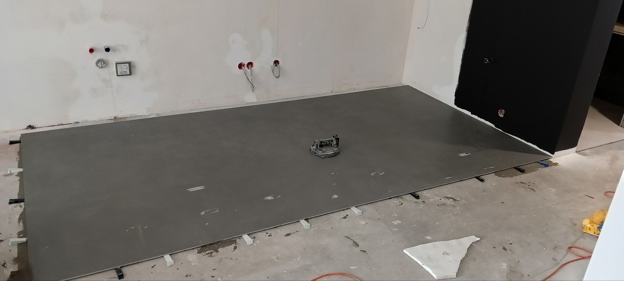 Wyrównana, szara wylewka samopoziomująca na podłodze w trakcie prac wykończeniowych. Widoczne przewody elektryczne i hydrauliczne w ścianie.