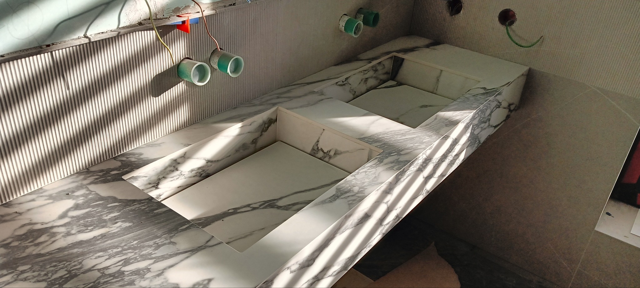 Biała umywalka z marmurowym blatem w trakcie montażu, widoczne rury i kable. Ściana wykończona w pionowe paski, oświetlona promieniami słońca.