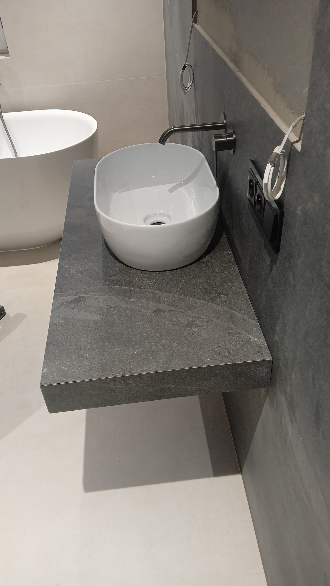 Nowoczesna łazienka z owalną umywalką nablatową na szarym blacie z kamienia, bateria ścienna w kolorze grafitowym, minimalistyczny design.