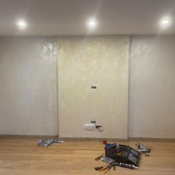 Pkartremonty - Pokój po szpachlowaniu z wykończoną ścianą w odcieniach beżu, drewnianą podłogą oraz narzędziami i puszkami elektrycznymi na podłodze, sugerującymi prace wykończeniowe.