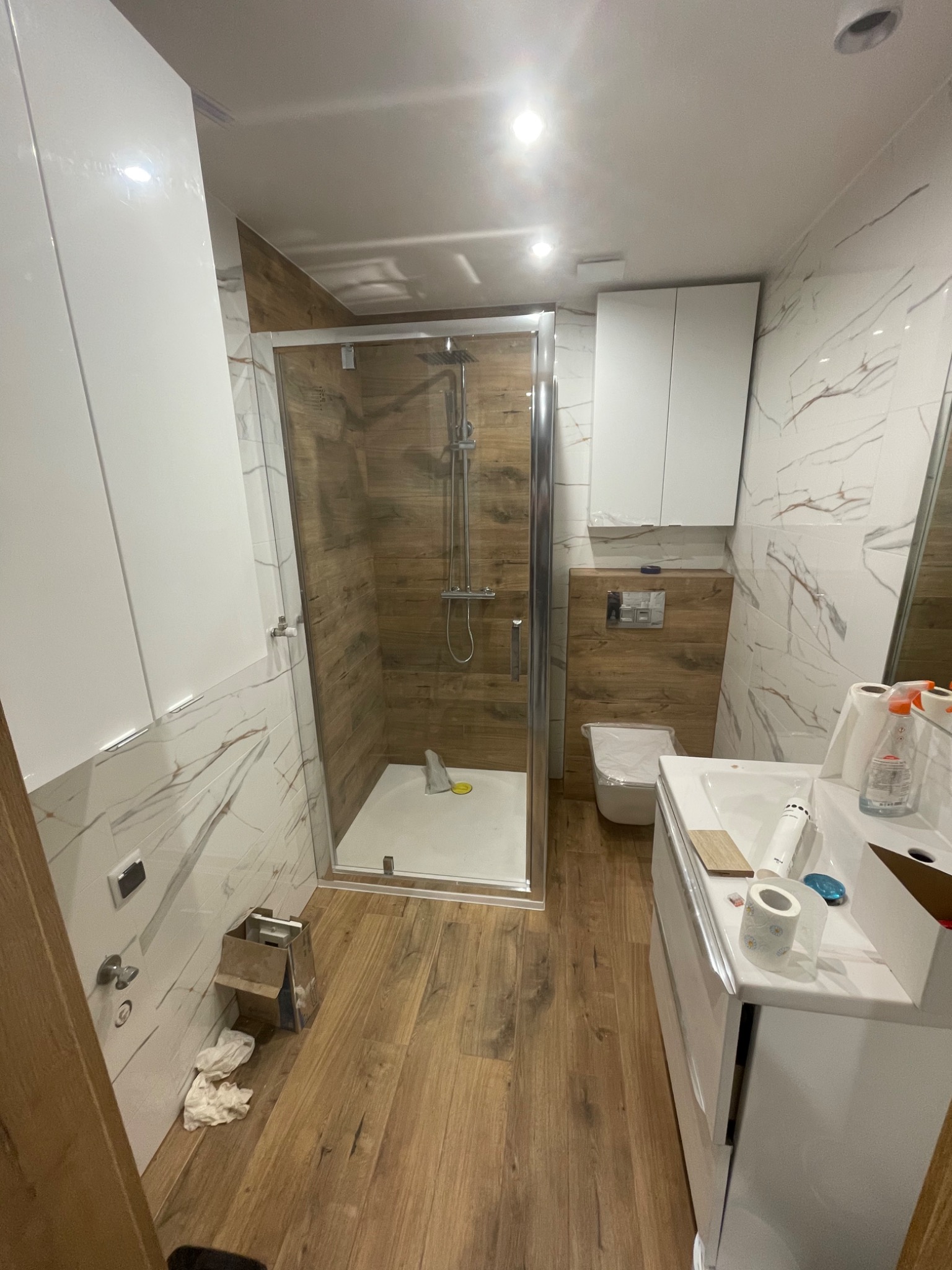 Nowoczesna łazienka po remoncie: kabina prysznicowa ze szklanymi drzwiami, białe szafki wiszące i umywalka, drewnopodobne płytki na podłodze i ścianie w kabinie.