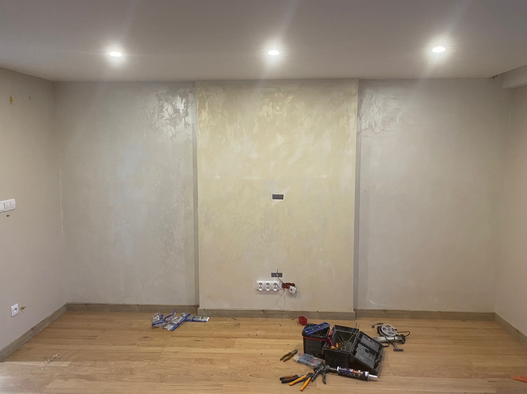 Pokój po szpachlowaniu z wykończoną ścianą w odcieniach beżu, drewnianą podłogą oraz narzędziami i puszkami elektrycznymi na podłodze, sugerującymi prace wykończeniowe.