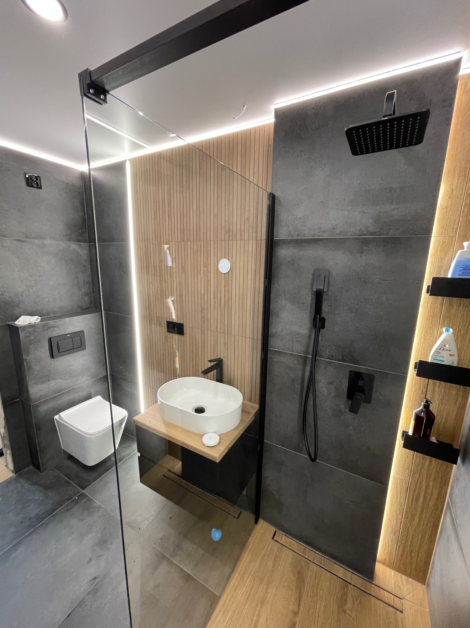 Nowoczesna łazienka z drewnianymi akcentami, szarą armaturą i kabiną prysznicową typu walk-in z odpływem liniowym. Umywalka nablatowa na drewnianym blacie.