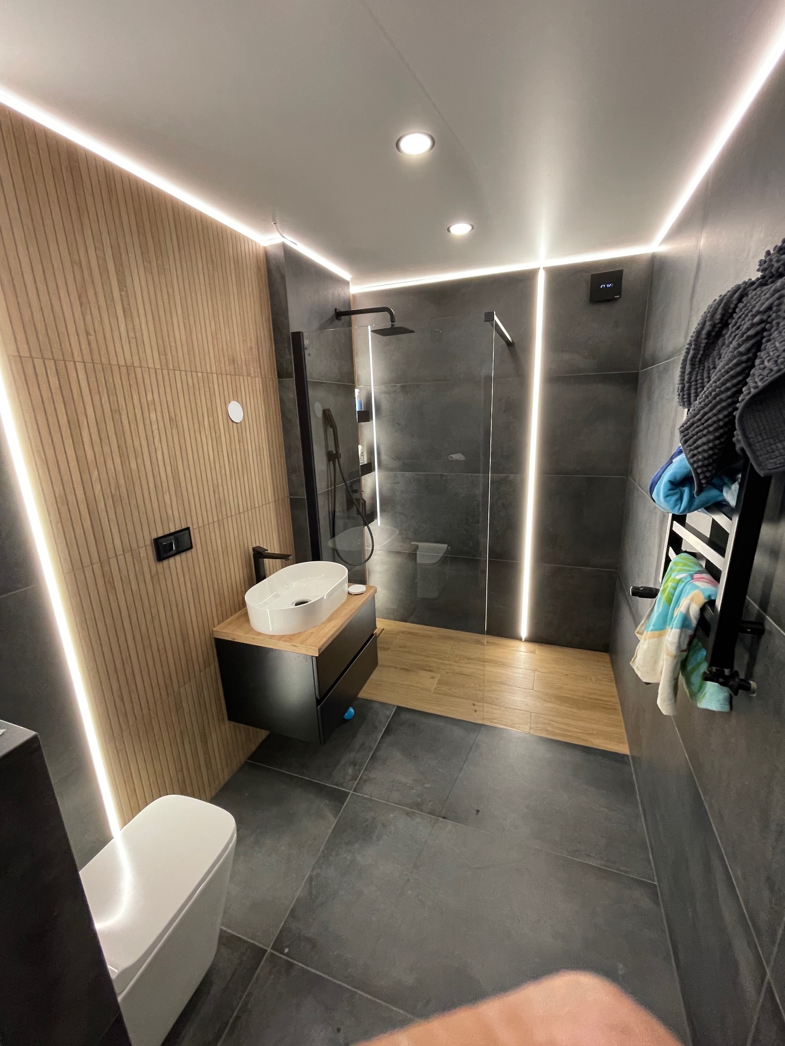 Nowoczesna łazienka z oświetleniem LED: drewniana ściana, czarne płytki, kabina prysznicowa z drewnianym podestem, umywalka nablatowa i wisząca toaleta.