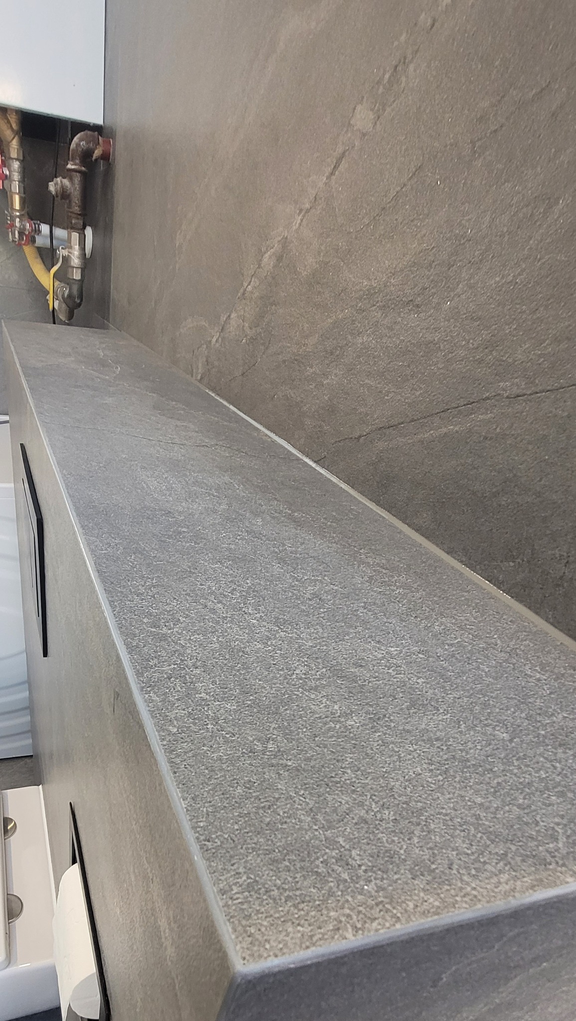 Wykończona ściana i blat w łazience z szarej, kamiennej płytki. Widoczne elementy instalacji. Minimalistyczny design.