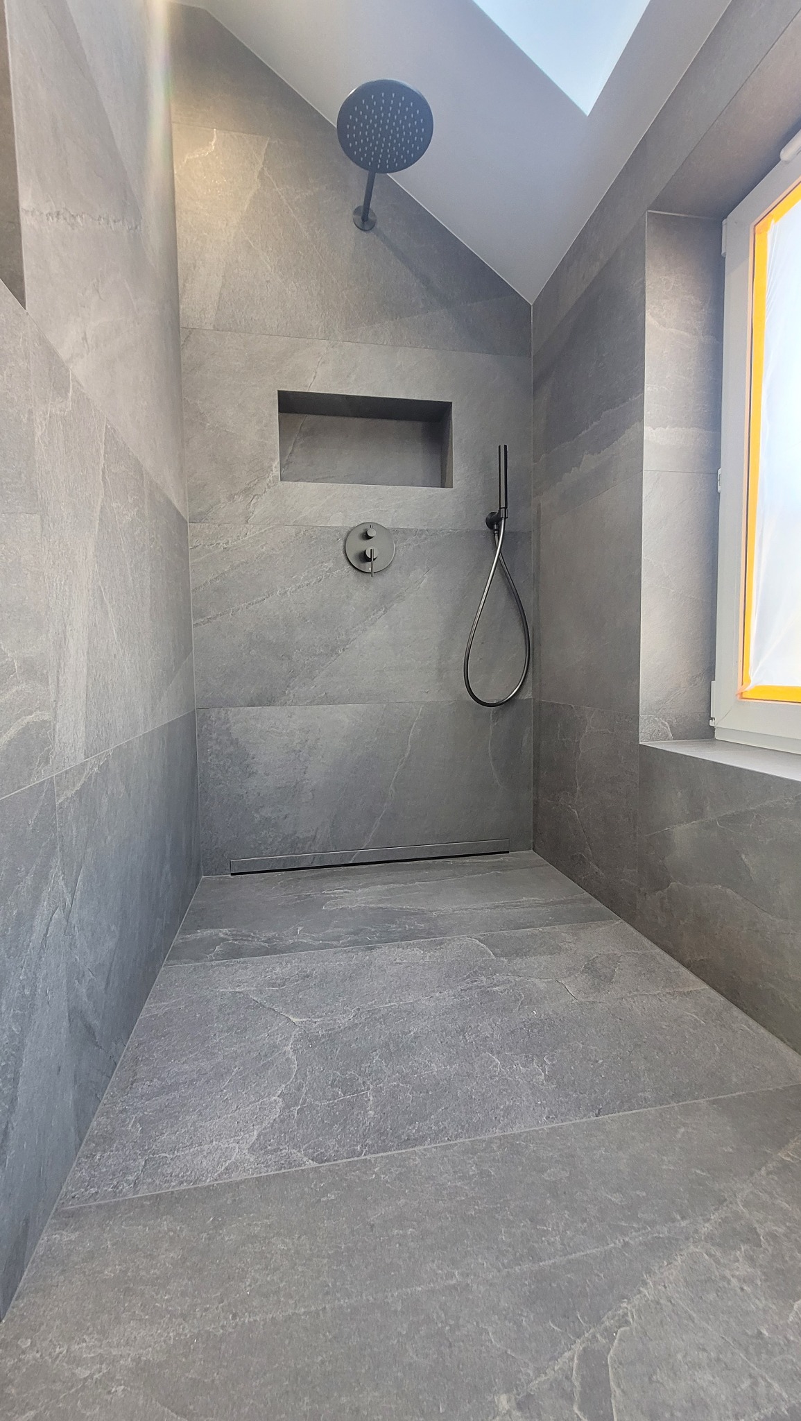 Wykończona kabina prysznicowa z szarymi płytkami imitującymi kamień, deszczownicą i wnęką w ścianie. Minimalistyczny design, nowoczesne wykończenie łazienki.