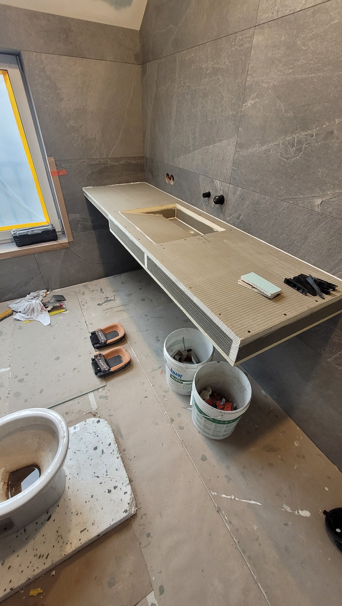 Wnęka umywalkowa z szarej płyty, obudowana mozaiką, w trakcie montażu, z widocznymi narzędziami i materiałami budowlanymi. Ściany wyłożone płytkami w podobnym odcieniu.