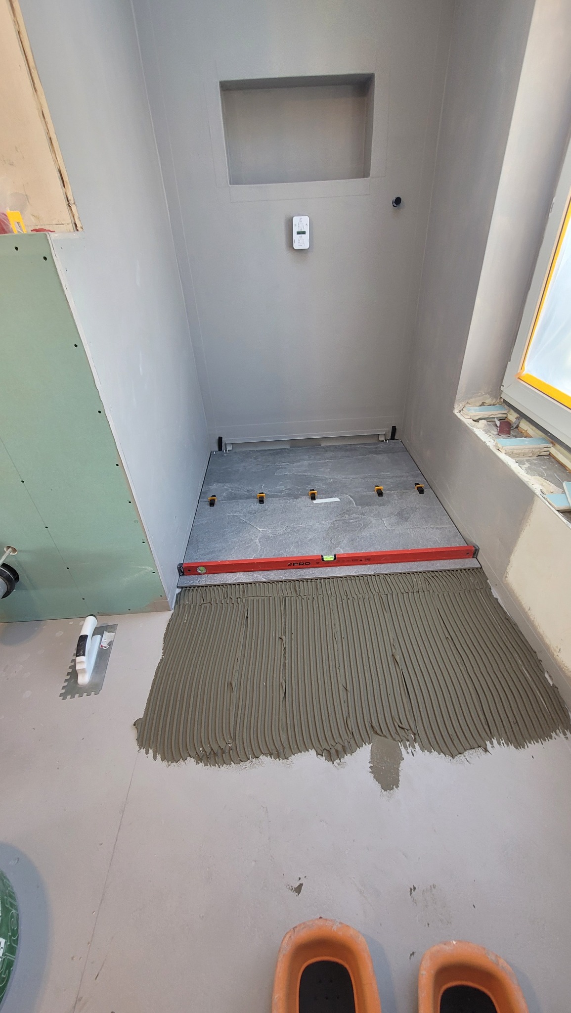 Układanie płytek podłogowych w łazience w Krakowie. Widoczna warstwa kleju, poziomica i fragment ułożonych płytek. Ściany przygotowane pod wykończenie.