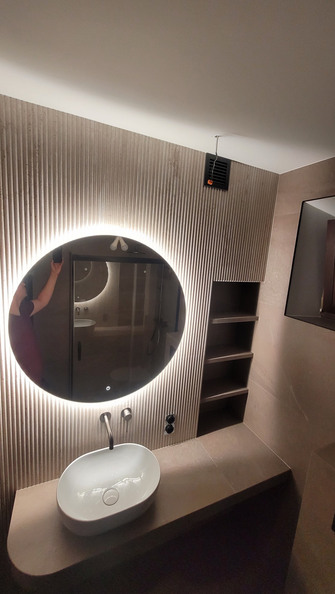 Nowoczesna łazienka z umywalką nablatową, podświetlanym lustrem i półkami w ścianie. Ściany wykończone w beżowych panelach z subtelnym wzorem. Minimalistyczny design.