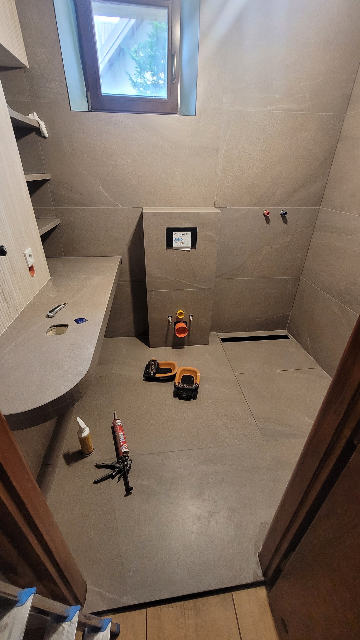 Wykończona łazienka z szarymi płytkami, widoczne narzędzia glazurnicze, zamontowana podtynkowa instalacja WC i odpływ liniowy. Ujęcie z poziomu podłogi.