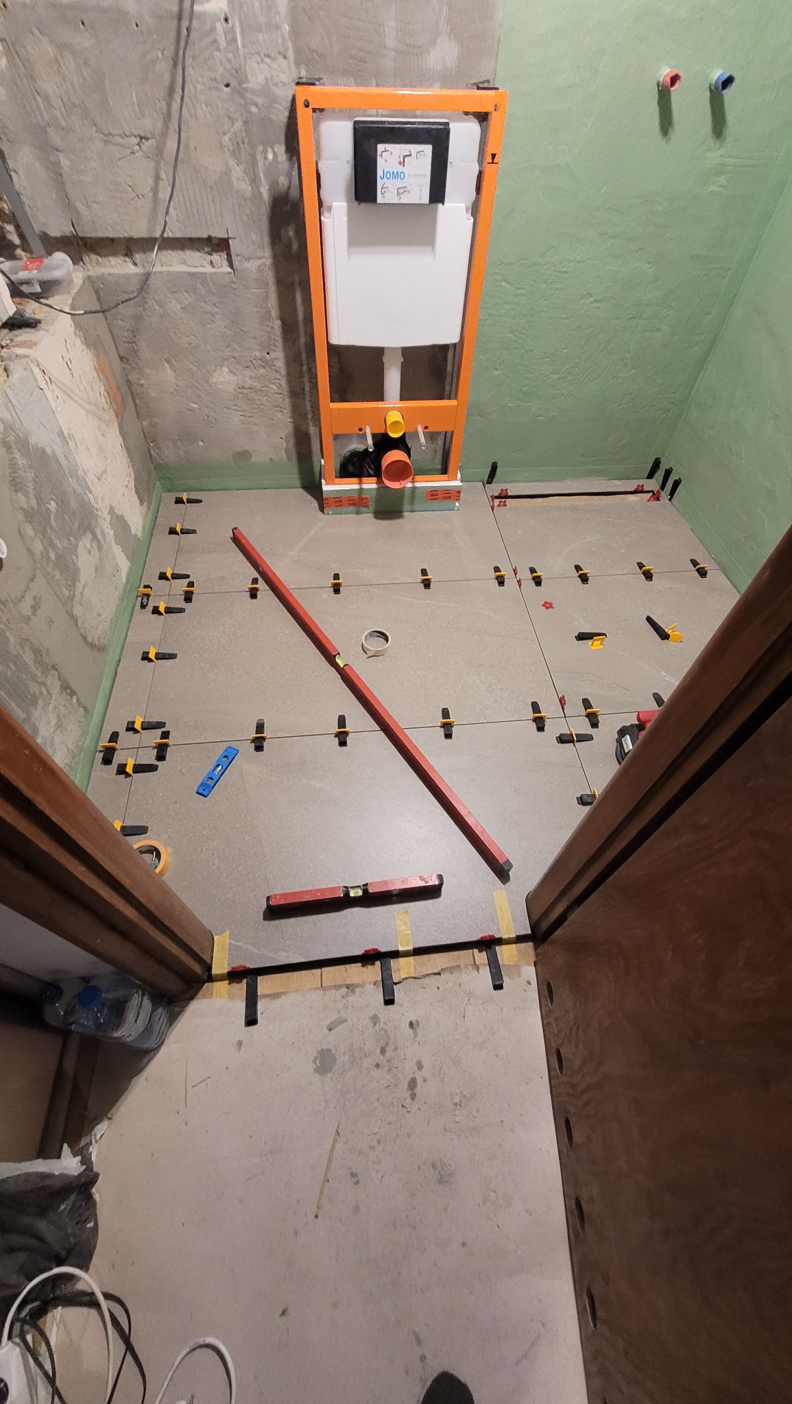 Montaż podtynkowego stelaża WC i układanie płytek podłogowych w łazience. Widoczny poziomica, krzyżyki dystansowe i przymocowany do ściany stelaż z instalacją.