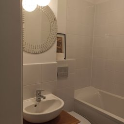 Twój Dom - Jasna łazienka z białą armaturą, okrągłym lustrem z mozaiką i drewnianą szafką pod umywalką. Widoczne białe płytki na ścianach i wanna.