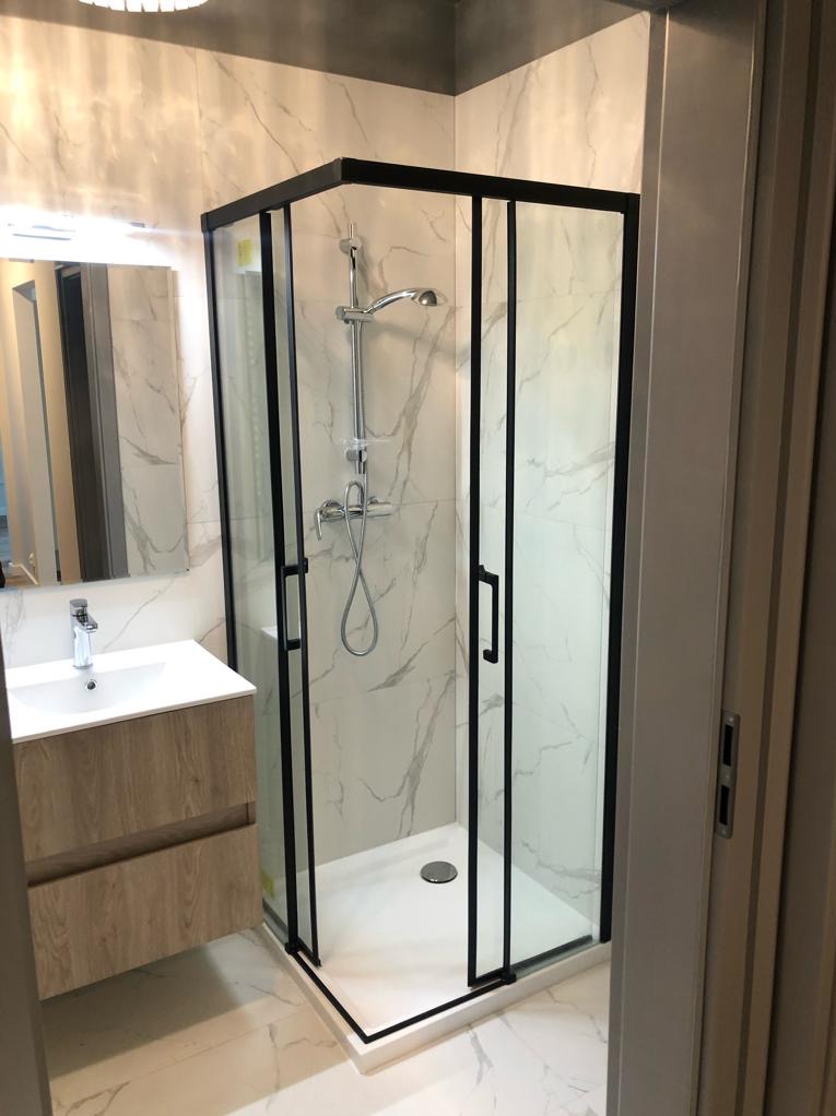 Nowoczesna kabina prysznicowa z czarnymi ramami i armaturą, marmurowe płytki na ścianach i podłodze, umywalka z szafką w jasnym drewnie. Minimalistyczny design.