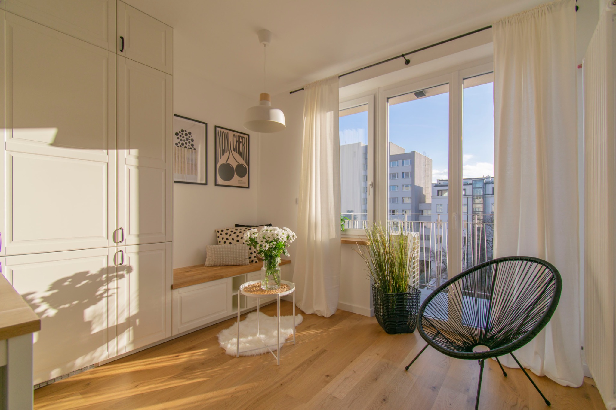 Jasne, nowoczesne wnętrze z zabudową szaf, siedziskiem przy oknie, okrągłym krzesłem i kwiatami w wazonie. Widok na budynki z balkonu.