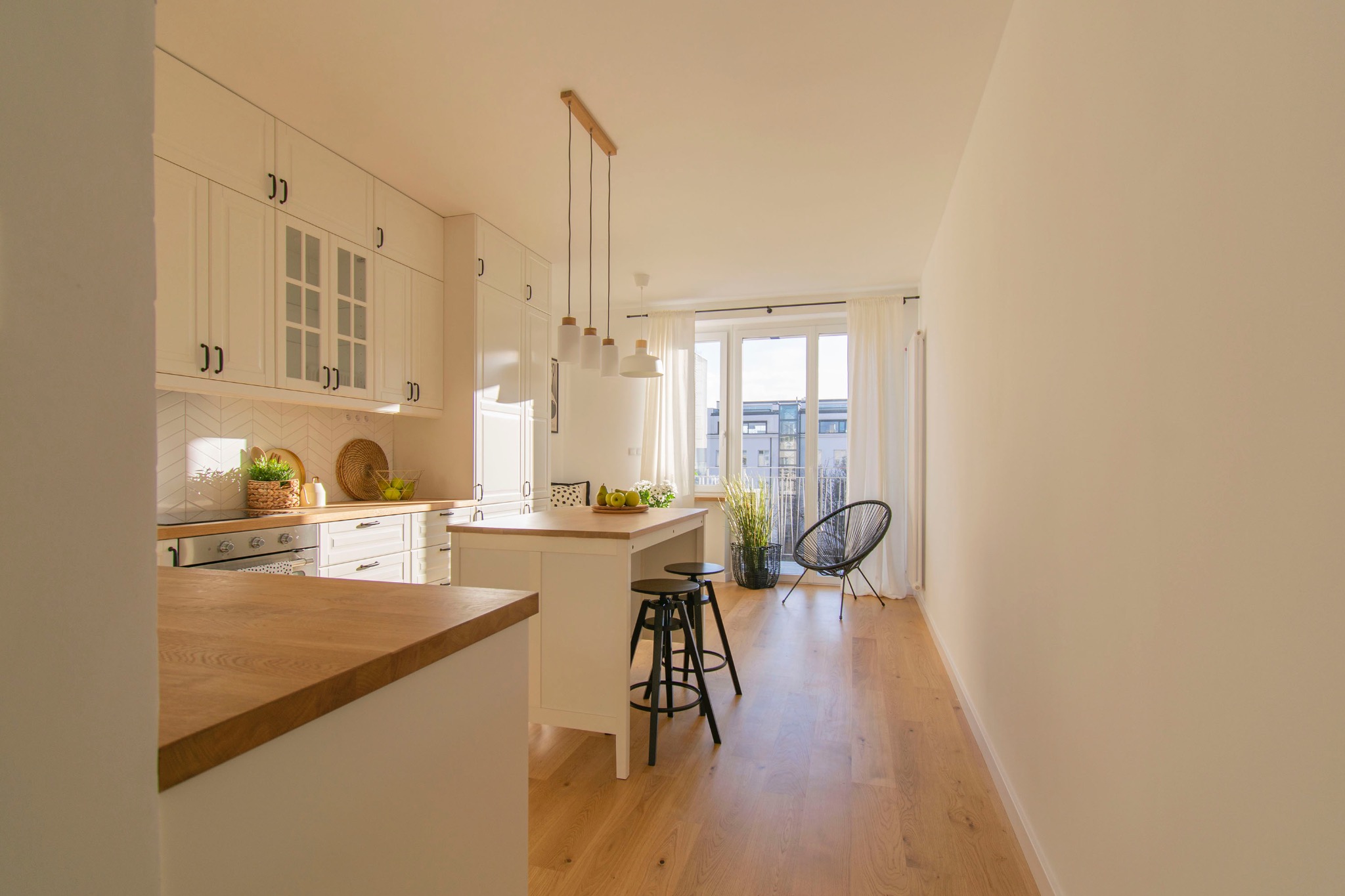 Jasna kuchnia z wyspą i drewnianym blatem, widok na balkon przez okno. Nowoczesne krzesła barowe i plecione krzesło na balkonie.