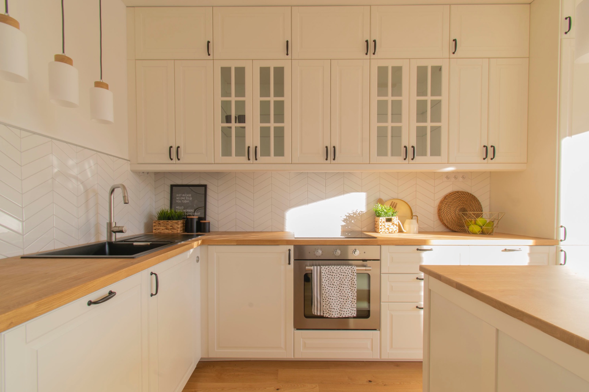 Jasna kuchnia w stylu skandynawskim: białe szafki z ciemnymi uchwytami, drewniany blat, chevronowe płytki. Oświetlenie punktowe i dekoracje. Porządek i estetyka.