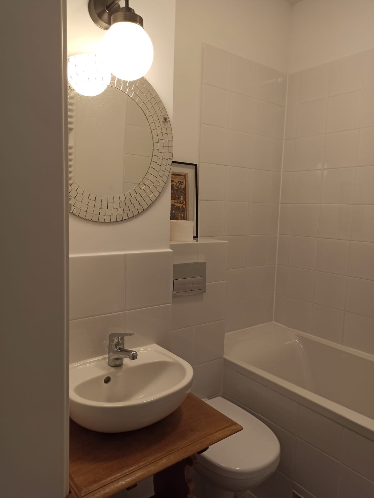 Jasna łazienka z białą armaturą, okrągłym lustrem z mozaiką i drewnianą szafką pod umywalką. Widoczne białe płytki na ścianach i wanna.