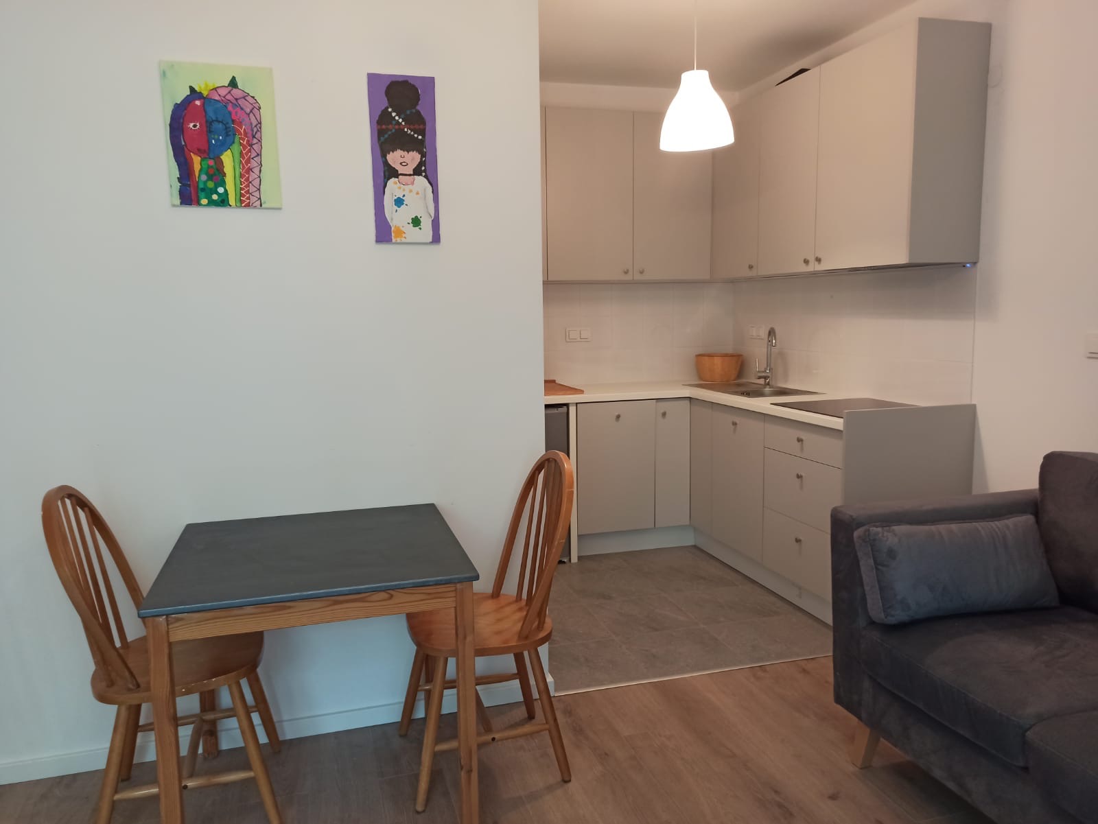 Jasna kuchnia z szarymi szafkami, drewnianym stołem i krzesłami. Na ścianie wiszą dwa kolorowe obrazy. Widoczny fragment szarej sofy.