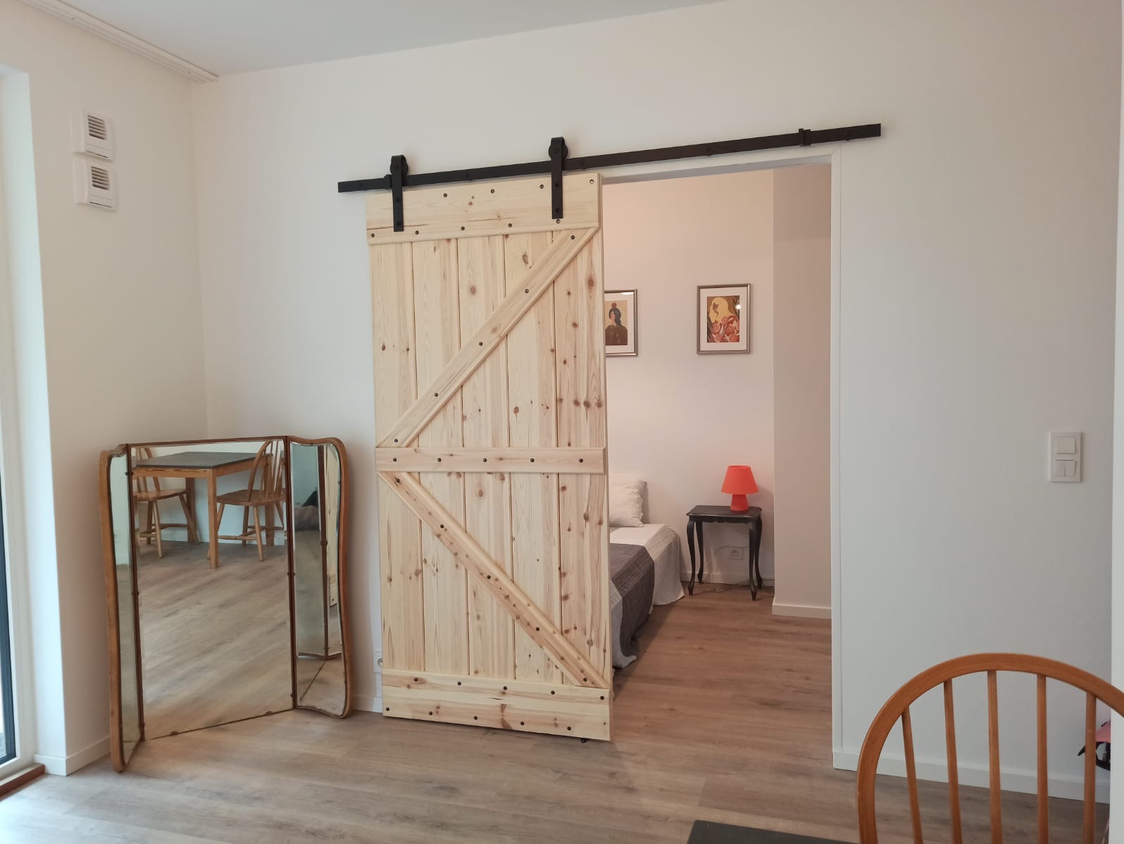 Drewniane drzwi przesuwne w stylu rustykalnym, zamontowane na czarnej szynie. W tle widoczne wnętrze pokoju z łóżkiem i stolikiem nocnym, lustro i podłoga z paneli.