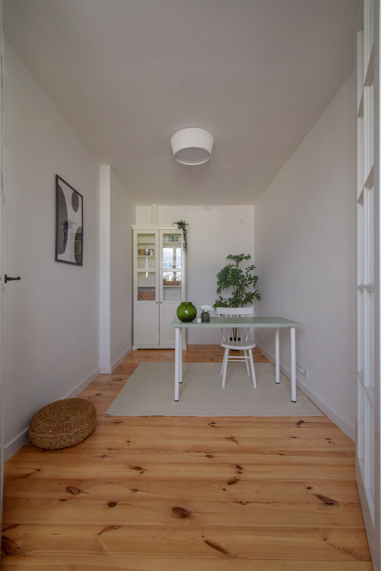 Jasne wnętrze z drewnianą podłogą: biurko, krzesło, szafka z przeszklonymi drzwiami i rośliny. Minimalistyczny wystrój, stonowana kolorystyka, dywan w paski i pleciony puf.