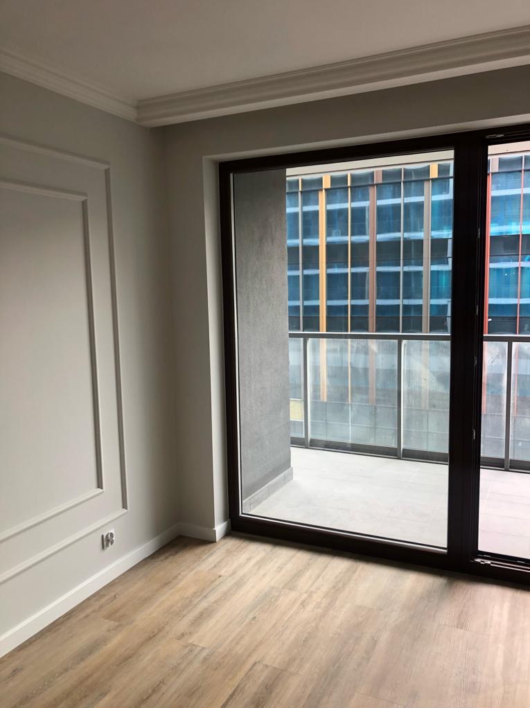 Jasne wnętrze z panelami podłogowymi i dekoracyjną sztukaterią ścienną. Widok na balkon przez duże okno z ciemną ramą, za którym widać nowoczesny budynek.