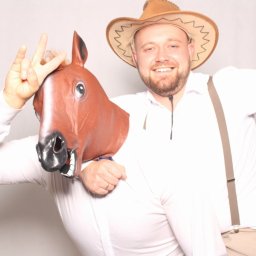 LuxEvent Joanna Kupiec - Uśmiechnięty mężczyzna w kowbojskim kapeluszu pozuje z osobą w masce konia, pokazując znak pokoju. Zabawna fotografia z imprezy, jasne tło.