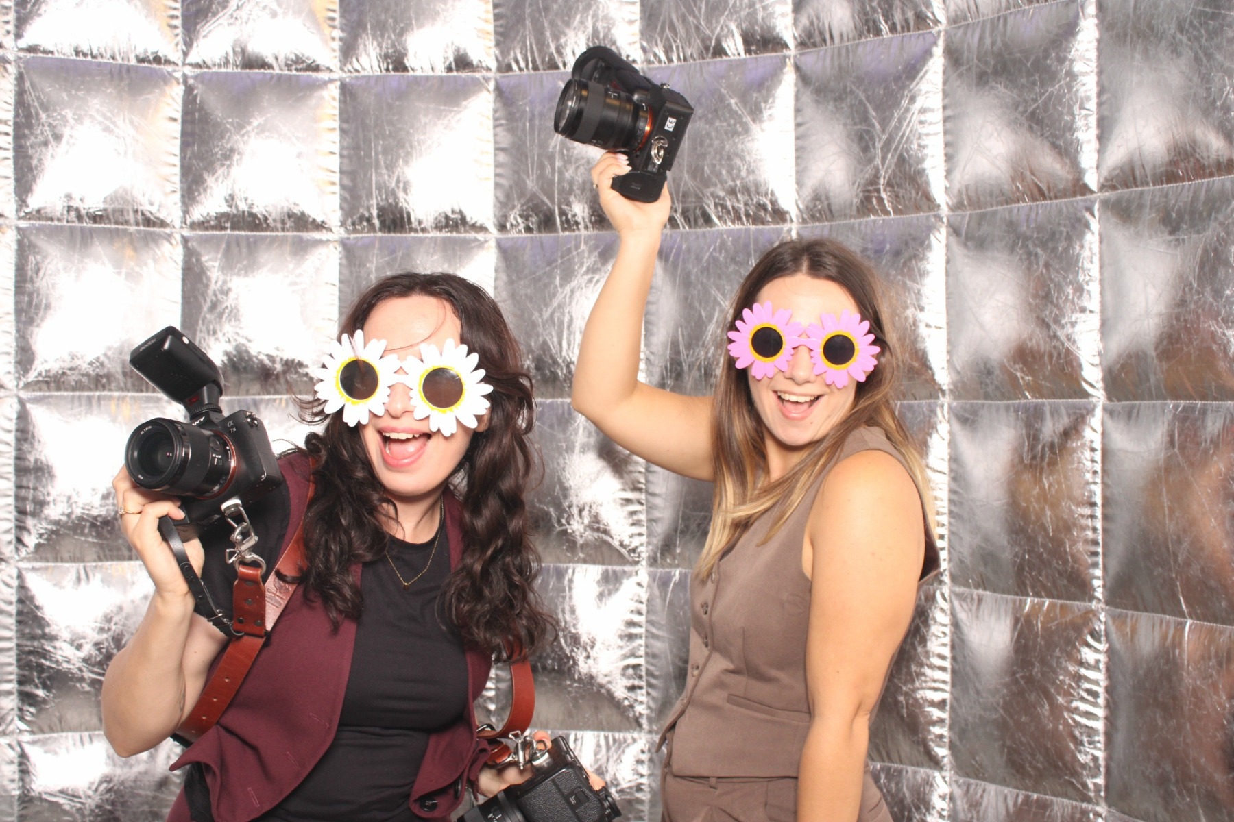 Dwie roześmiane kobiety w okularach w kształcie kwiatów, trzymające aparaty na tle srebrnej fotobudki. Zabawa i radość podczas imprezy.